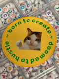 A round yellow sticker with a cat cutout in the center and green text reading 'born to create ~ doomed to hustle ~' is placed over a tray of white alphabet beads with colorful letters.