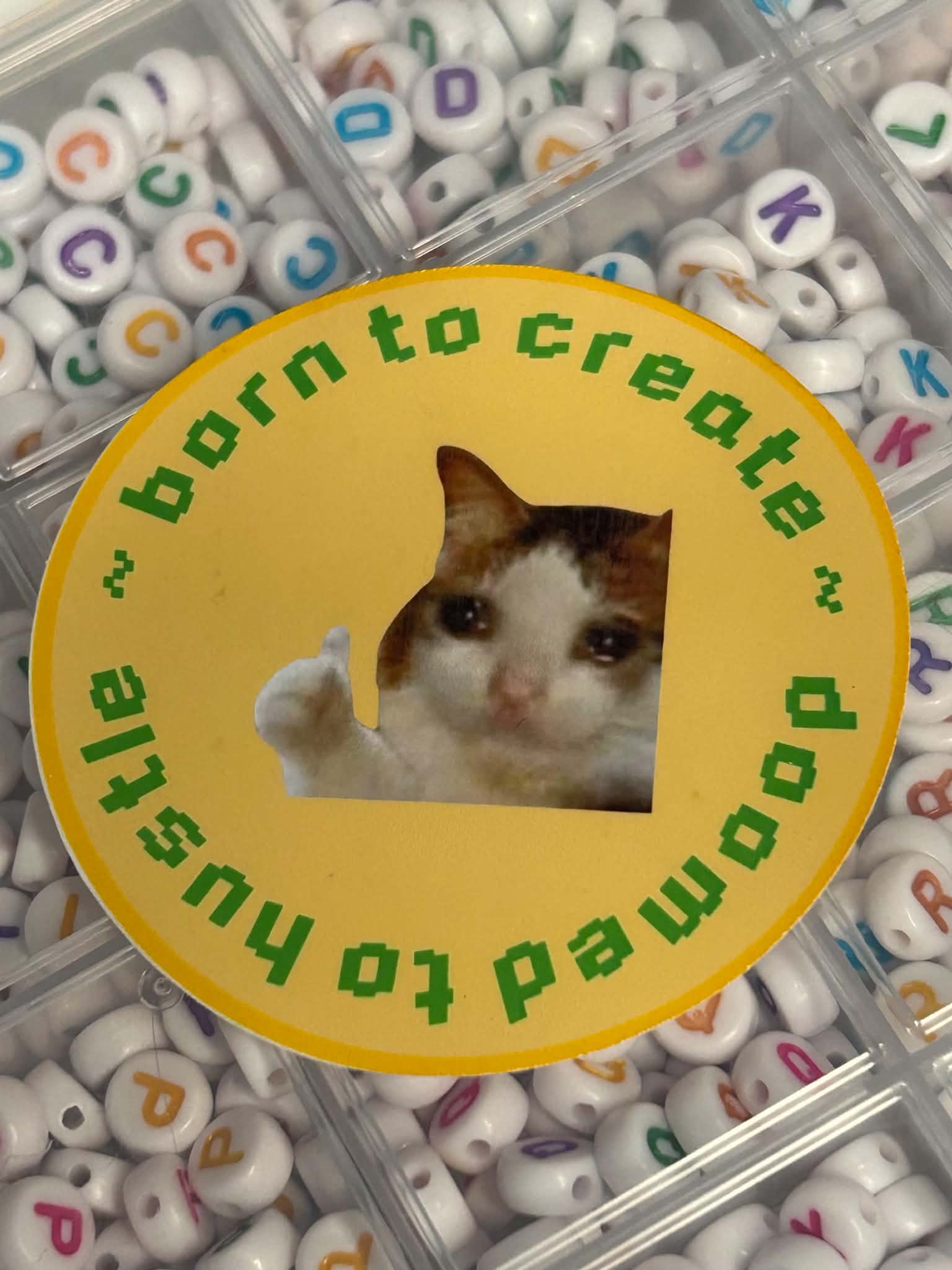 A round yellow sticker with a cat cutout in the center and green text reading 'born to create ~ doomed to hustle ~' is placed over a tray of white alphabet beads with colorful letters.