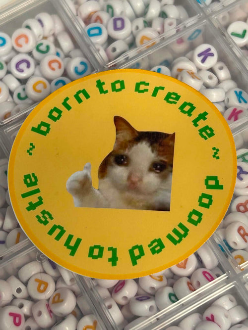 A round yellow sticker with a cat cutout in the center and green text reading 'born to create ~ doomed to hustle ~' is placed over a tray of white alphabet beads with colorful letters.